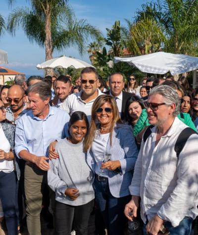 Gruppo di persone sorridenti all'aperto con palme e ombrelloni sullo sfondo.