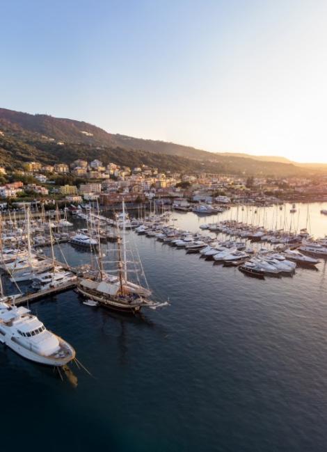 Porto turistico al tramonto con barche a vela e yacht, circondato da colline.