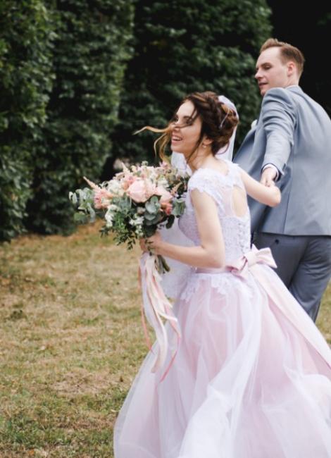 Sposi felici corrono nel parco, lei con bouquet e abito bianco e rosa.