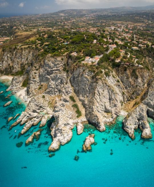 Scogliera imponente con acque turchesi e villaggio costiero immerso nel verde.